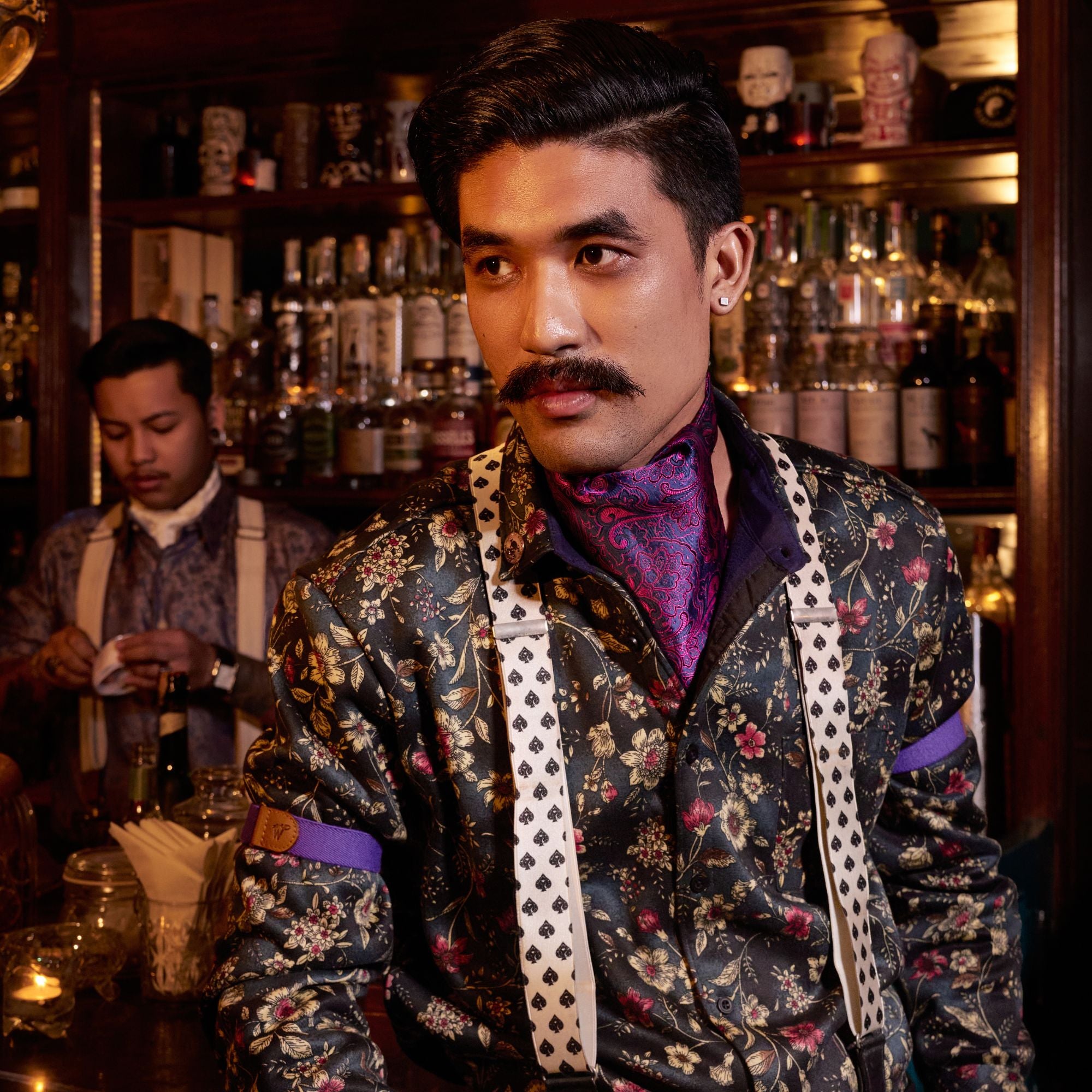 Person wearing patterned floral shirt, with suspenders with a spades pattern. He standing in front of a bar.