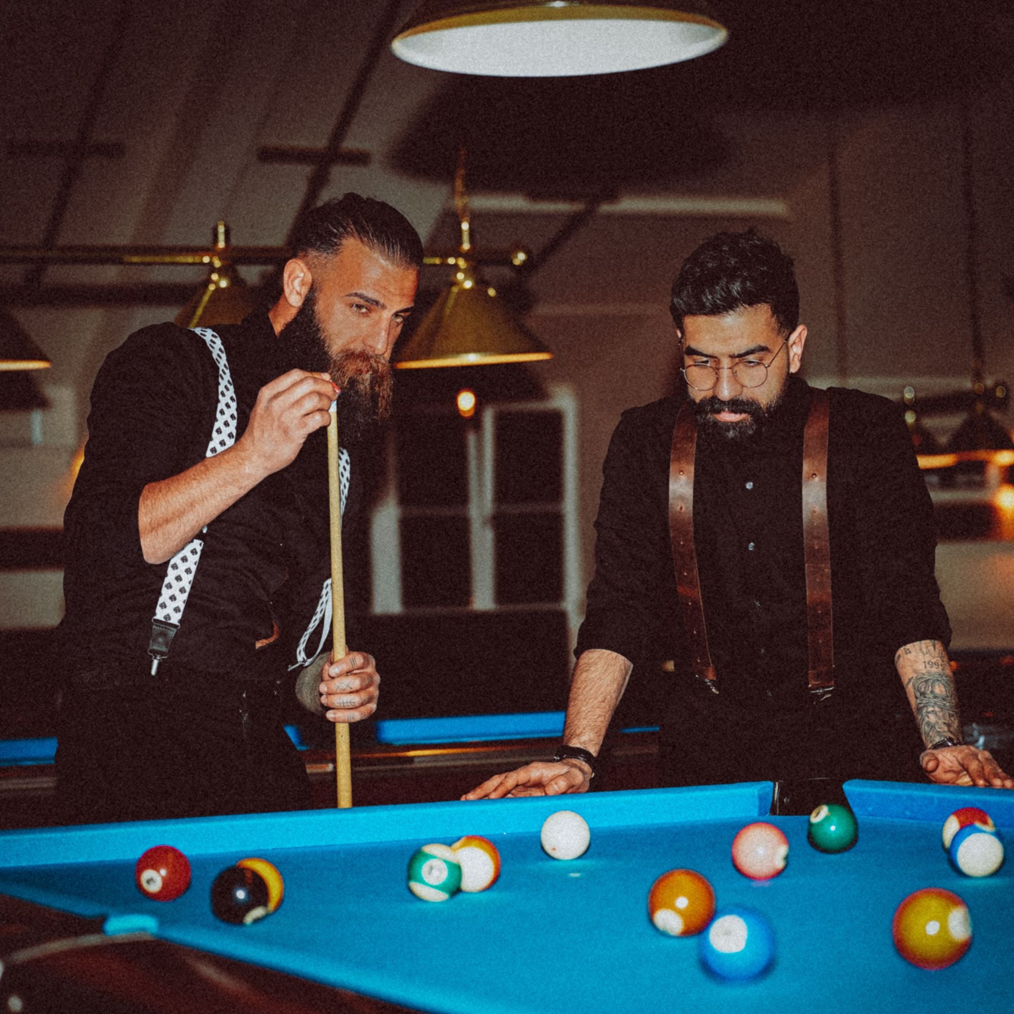 Two people playing pool at a blue pool table, both are wearing suspenders.