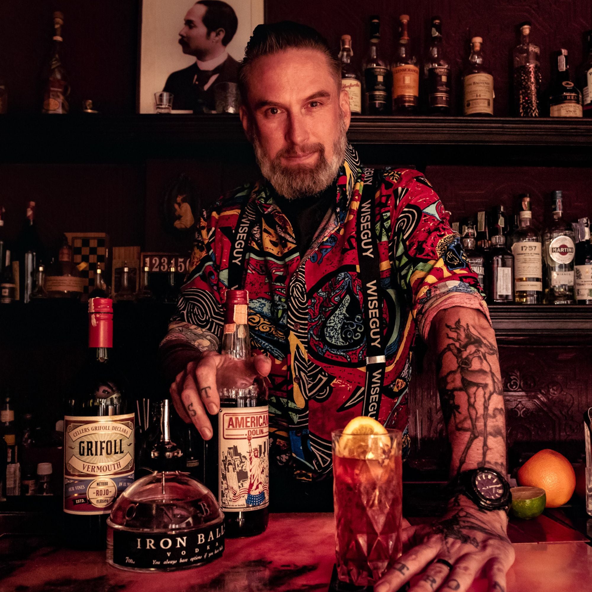Man who is bartender with rolled up sleeves showing tattoos. He is wearing elastic suspenders with the word 'Wiseguy' on them.