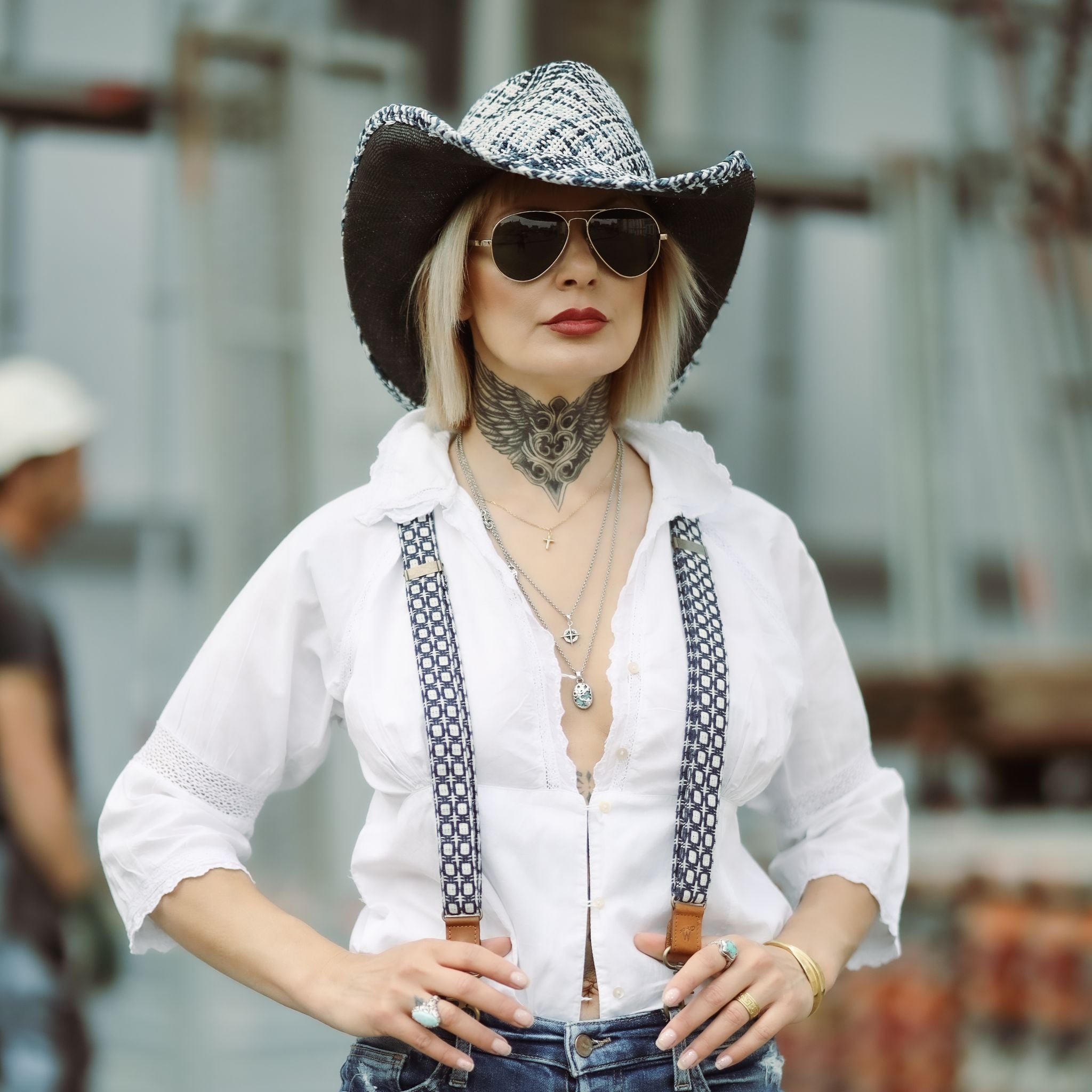 Woman wearing a blue and white cowboyhat and aviator sunglasses. She is wearing a white blouse and blue and white Suspenders.