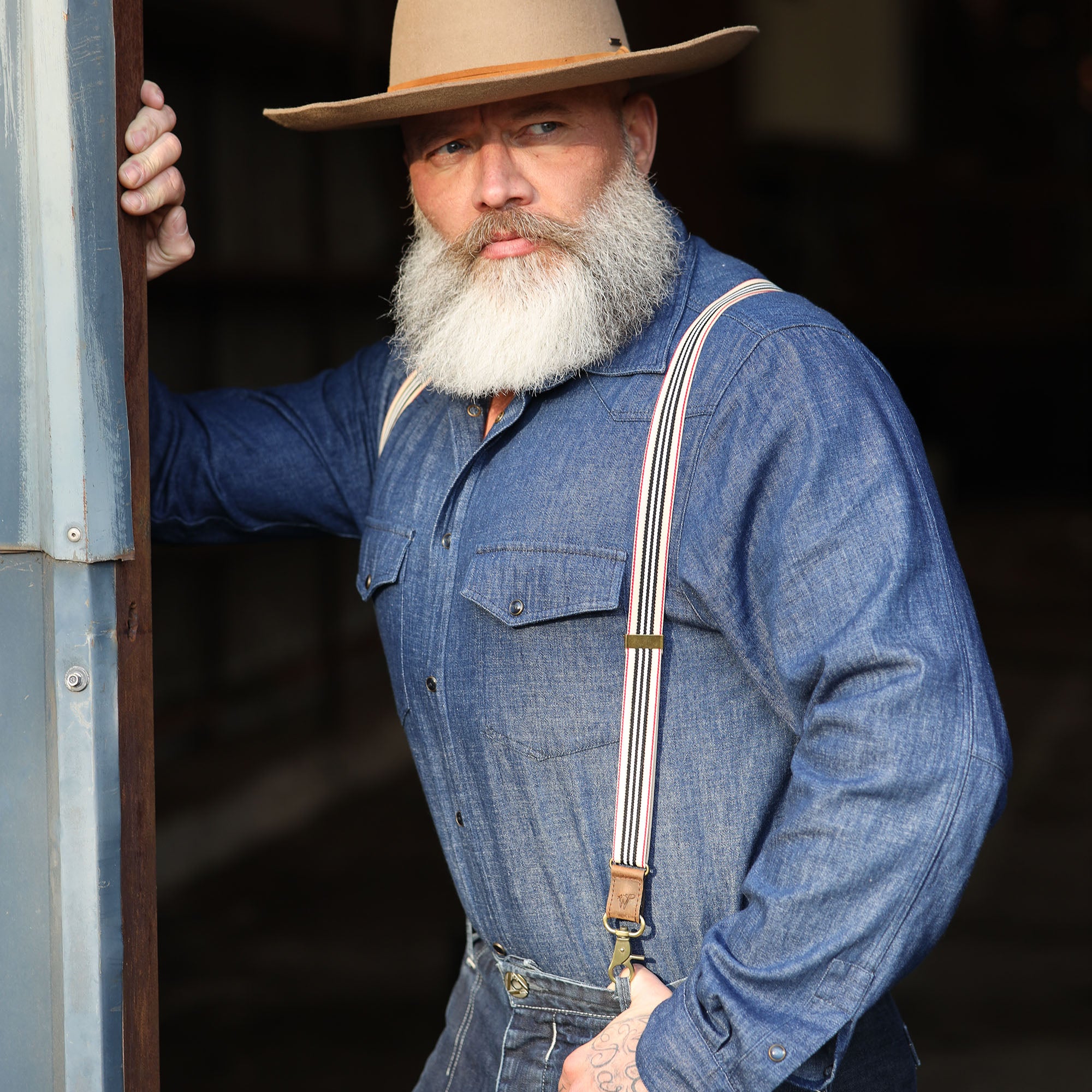 Man wearing Striped Wiseguy Racing Suspenders overa Denim Shirt, styled with a hat in a factory setting