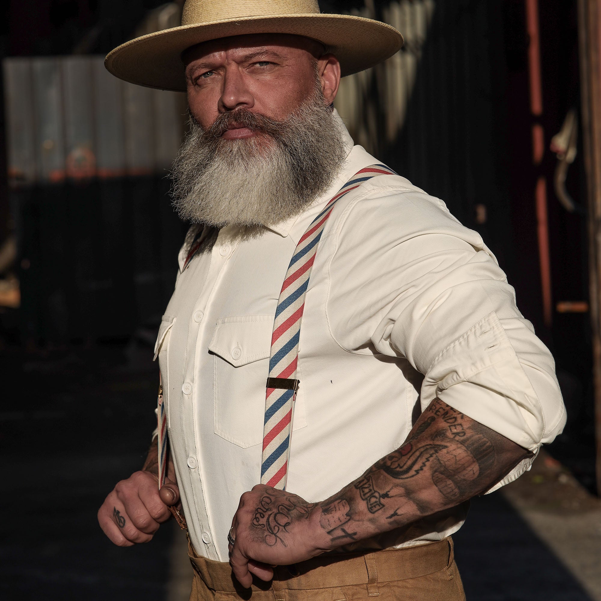 Man wearing Barber Pole striped Wiseguy Essential Suspenders over a white shirt, styled with a hat in a factory setting.