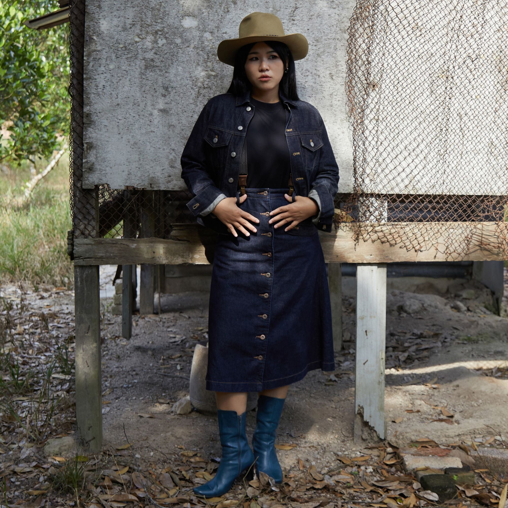 Mina wearing a cowboy hat, denim jacket, denim skirt, blue leather boots and Wiseguy Original herringbone pattern elastic suspenders.