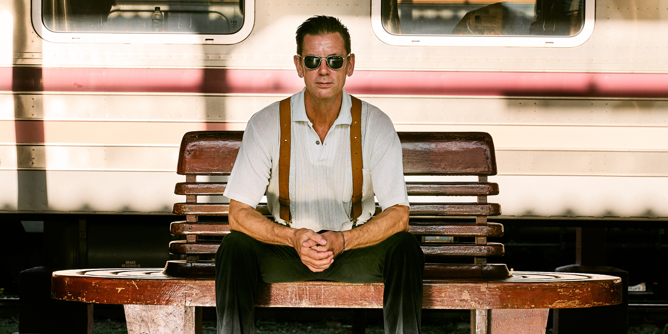 A man facing the camera, sitting on a bench at a train station. He is wearing aviator sunglasses and leather Wiseguy Original Crazy Horse Suspenders. There is a train behind him standing still.