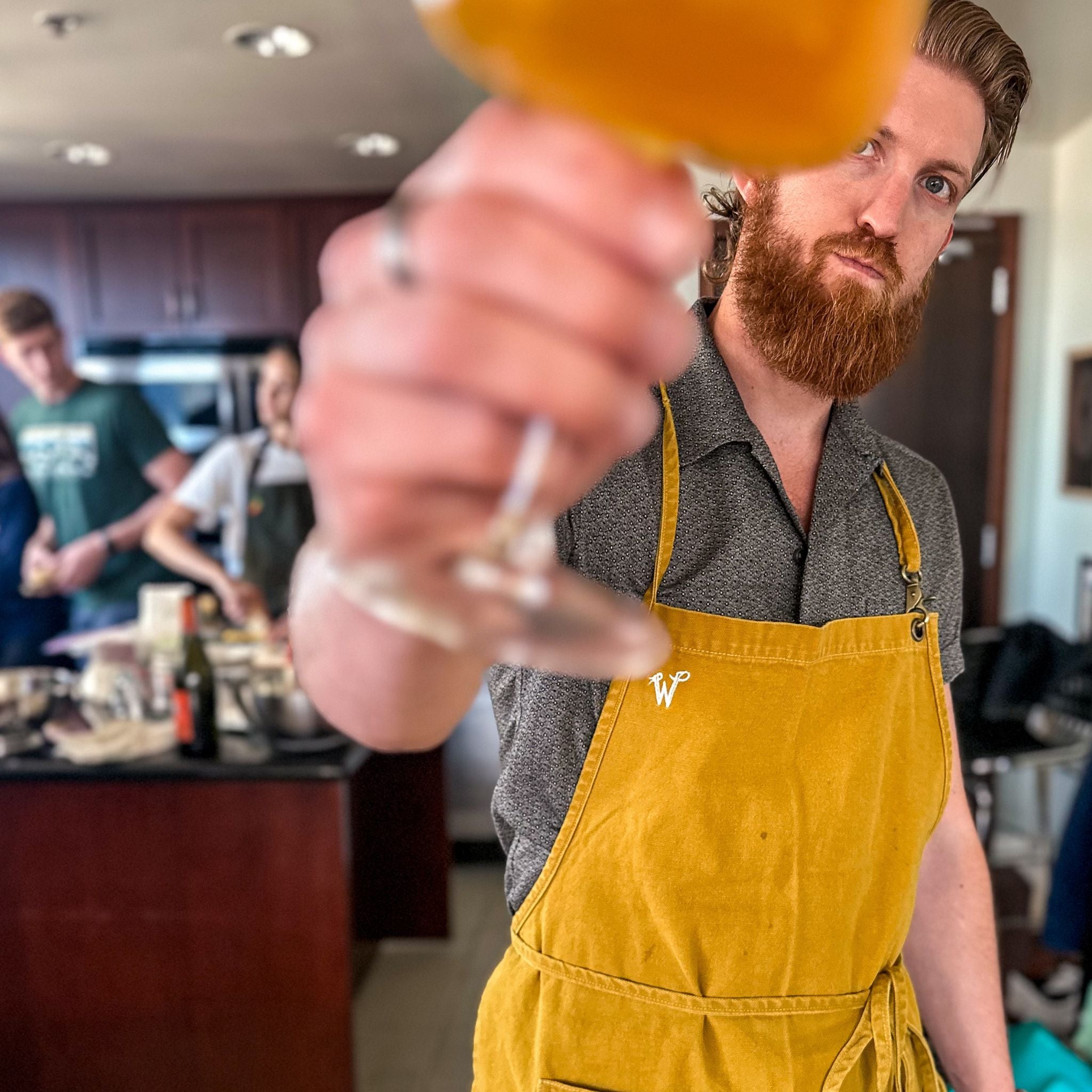 Cal is wearing the Wiseguy Original Apron in the color mustard, he is standing in the kitchen with a glass. 