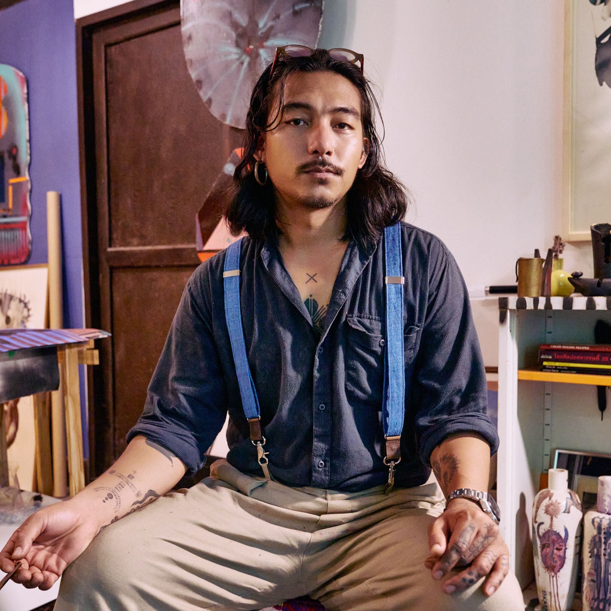 Man sitting down in art studio, looking into the camera. He's wearing a dark blue shirt and bright blue Suspenders.